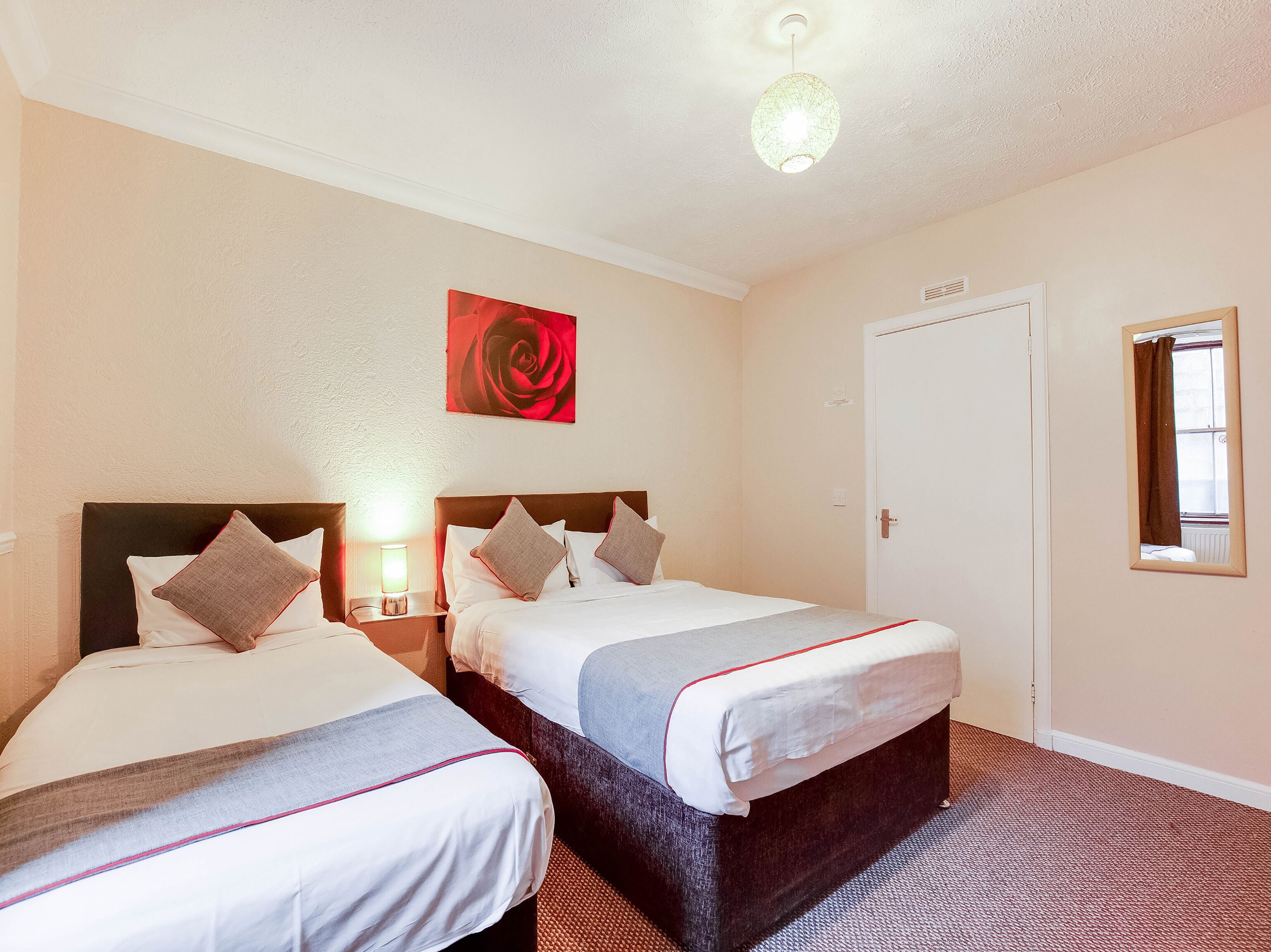 A hotel room with two beds, a red rose painting, a ceiling light, and a window with a curtain.