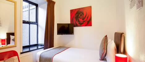 A hotel room with a bed, a red chair, a window with curtains, a wall-mounted TV, and a red rose painting.
