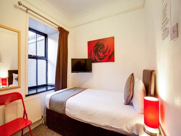 Habitación de hotel con una cama, una silla roja, una ventana con cortinas, un televisor montado en la pared y un cuadro de una rosa roja.
