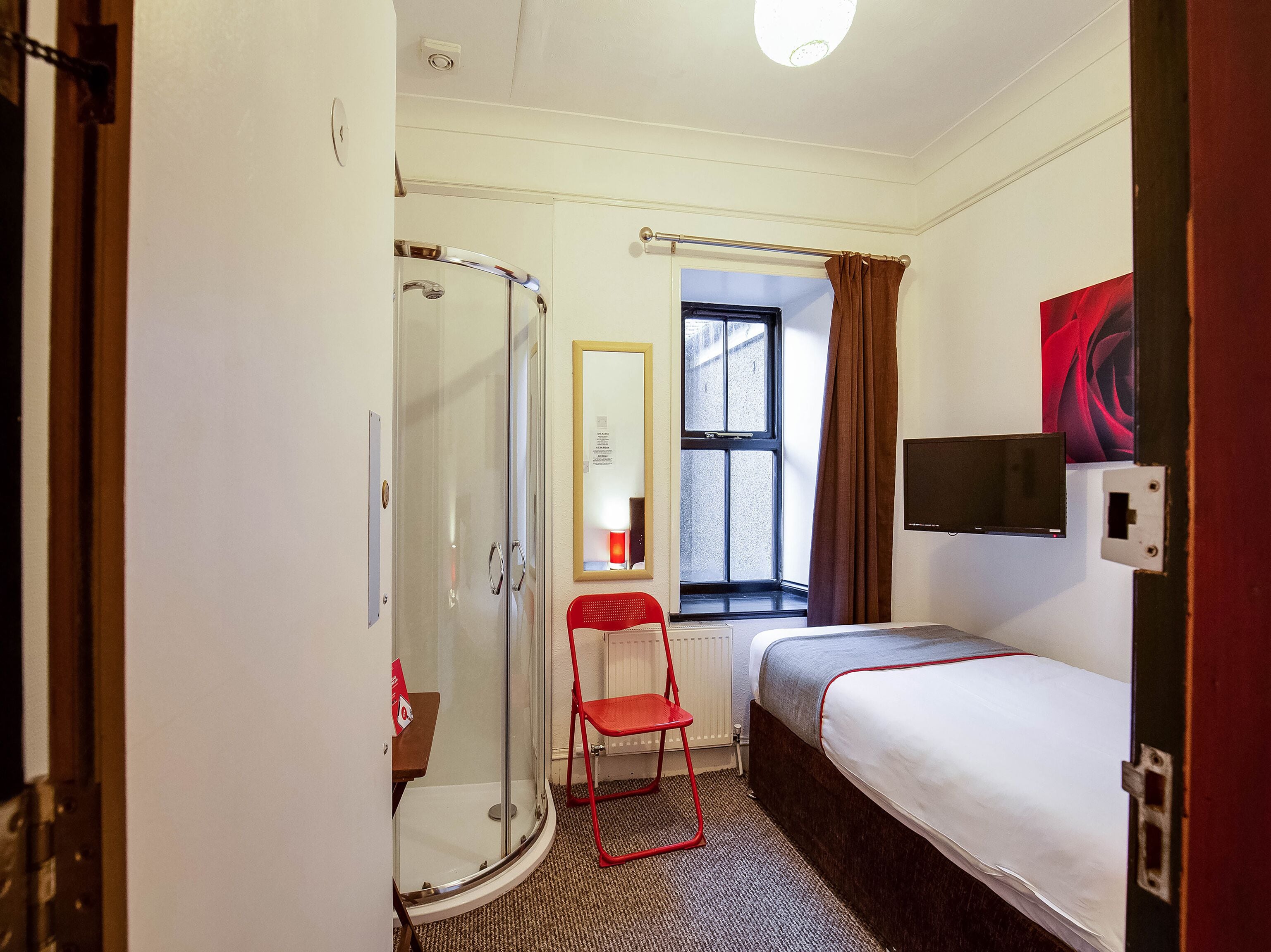 Une petite chambre d’hôtel avec une douche, un lit simple, une chaise et une fenêtre avec des rideaux.