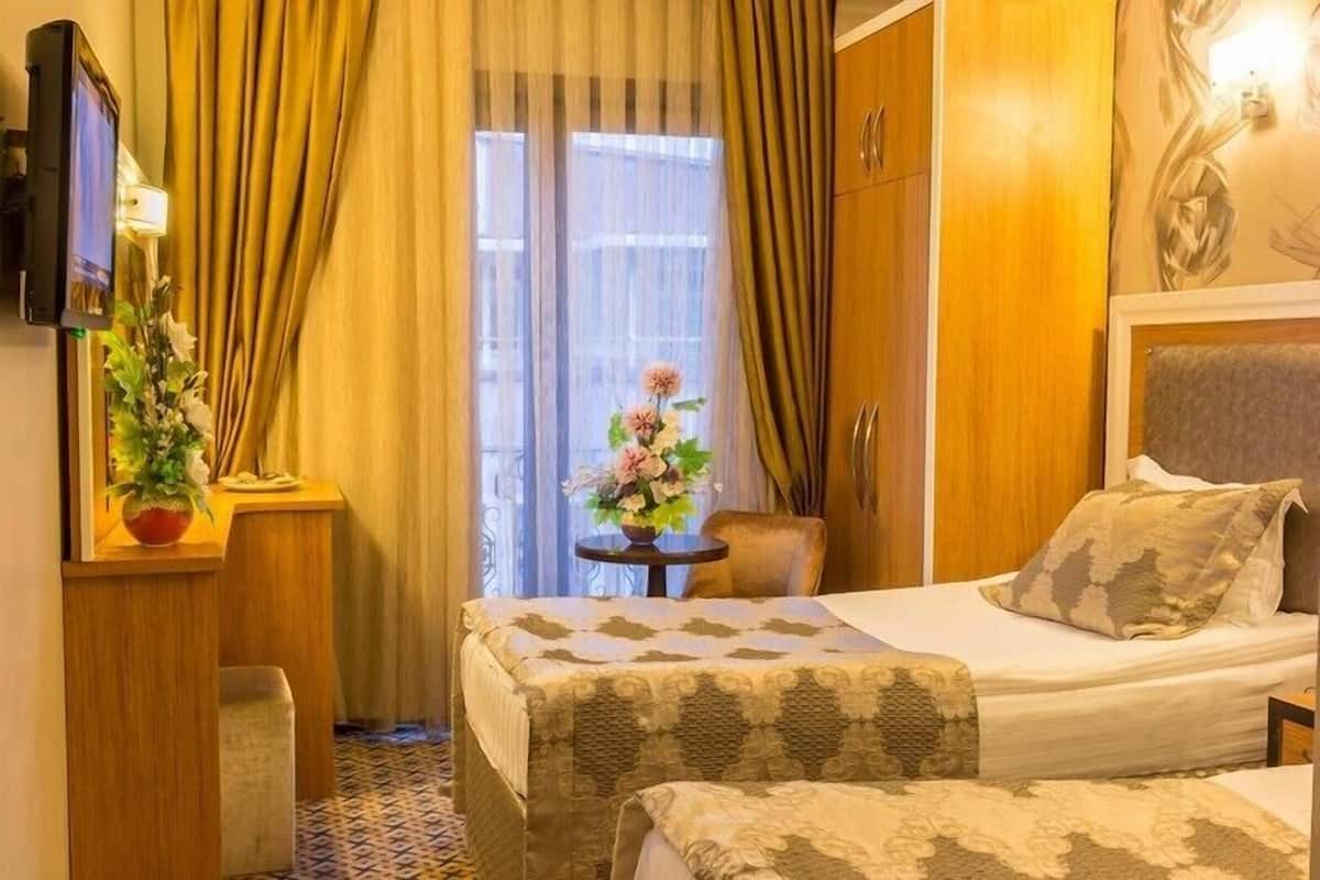 A hotel room with a bed, a TV, a small table with a vase of flowers, and a view of the outdoors through a window with curtains.