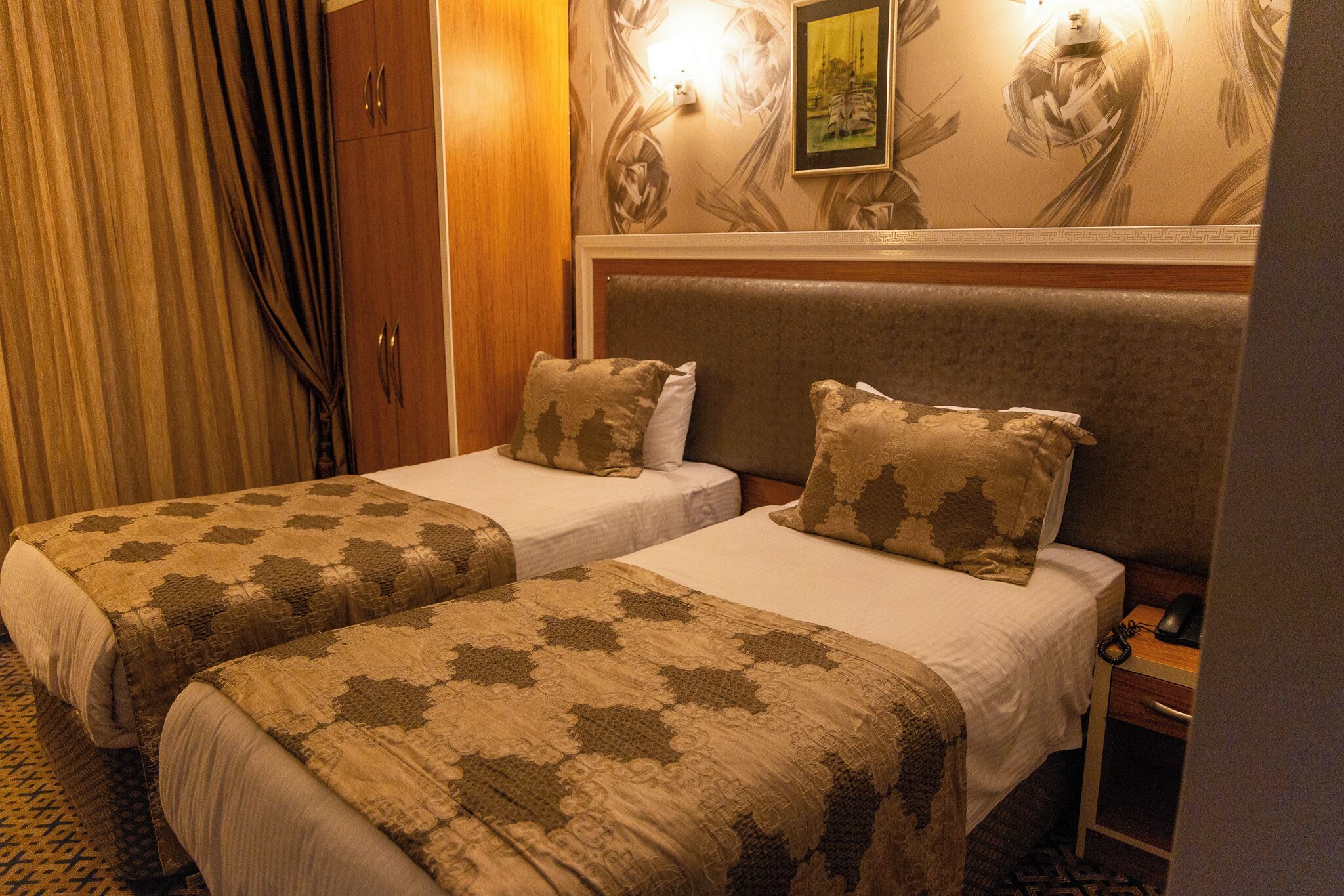 Ein Hotelzimmer mit zwei Betten, beide mit gemusterten Bettbezügen und Kissen. Ein Holzschrank und ein Nachttisch mit Lampe sind vorhanden.