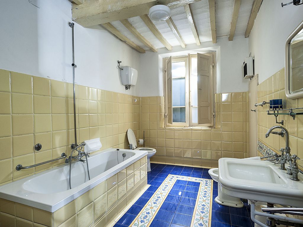 A bathroom with a bathtub, toilet, and sink, featuring yellow and blue tiles, wooden beams, and a window.