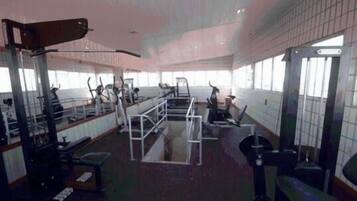 Un gimnasio con diverso equipo de ejercicio, como cintas de correr y máquinas de pesas, dispuesto en una sala amplia con ventanas grandes.