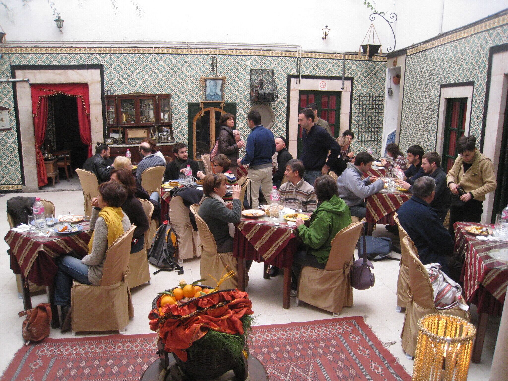 A crowded dining area with tables and chairs, a basket of oranges, and patterned walls.