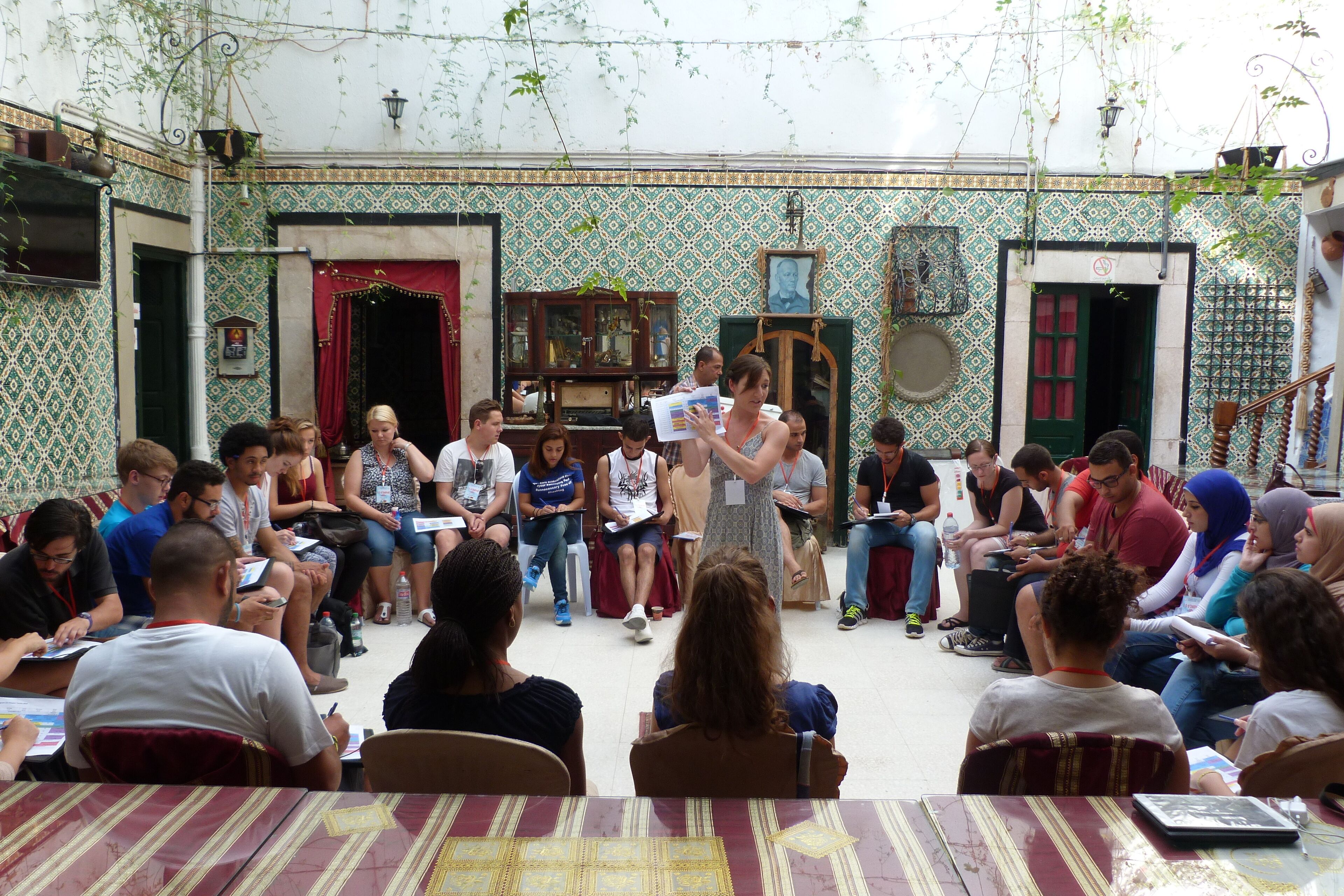 A group of people seated in a courtyard with patterned walls and a central area with a speaker.