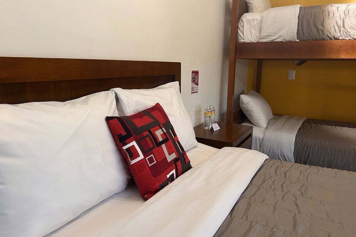 Habitación de hotel con una cama individual, una litera, una mesita de noche con una botella de agua y un cojín rojo estampado.