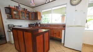A kitchen with wooden cabinets, a white refrigerator, and a window with a view of greenery. - Jaco Villa 42 (León XIII)