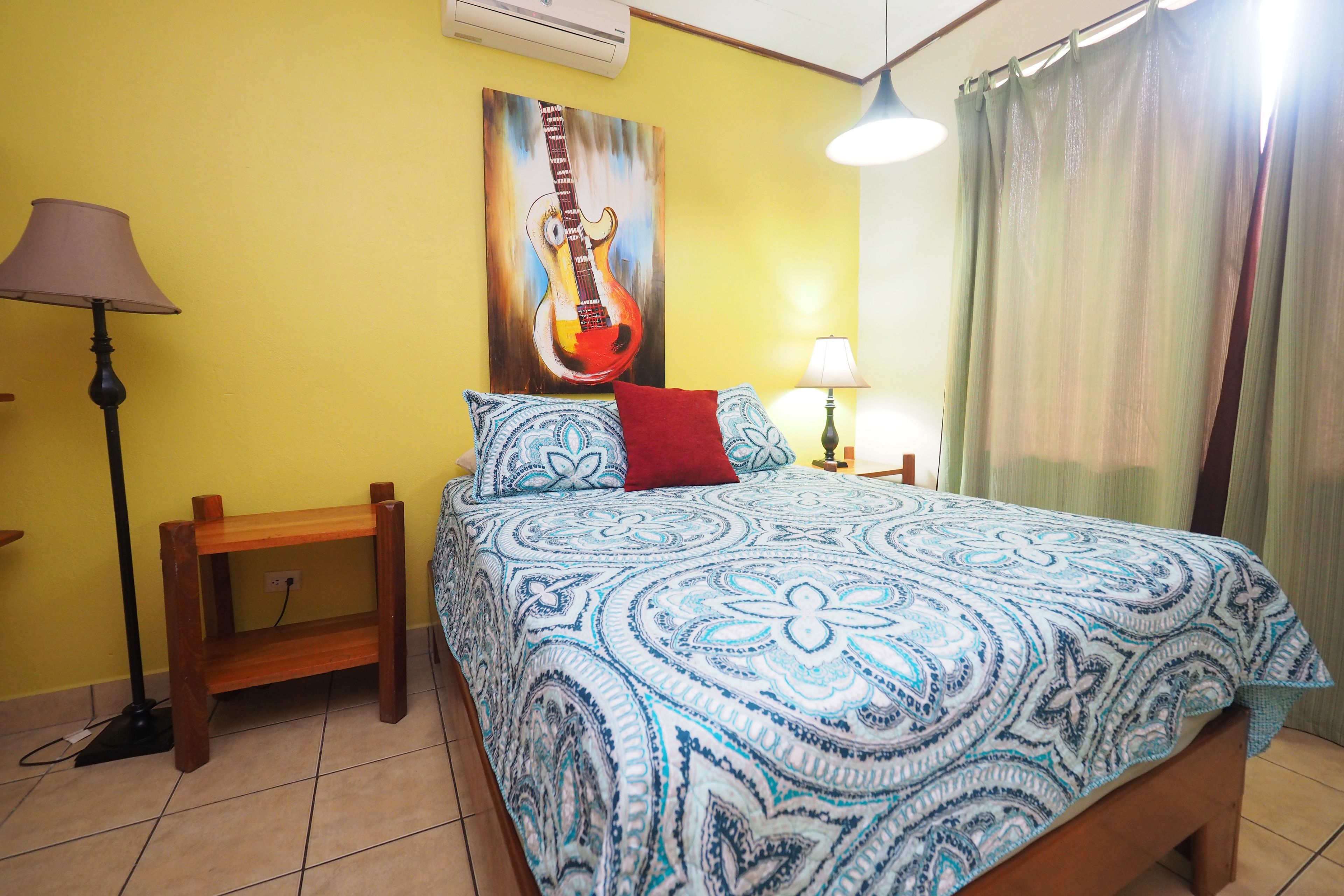 A bedroom with a patterned bedspread, a guitar painting on the wall, a wooden nightstand, and a lamp.