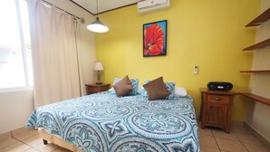 A bedroom with a bed, a nightstand, a lamp, a wall-mounted air conditioner, and a painting. - Jaco Villa 42 (León XIII)