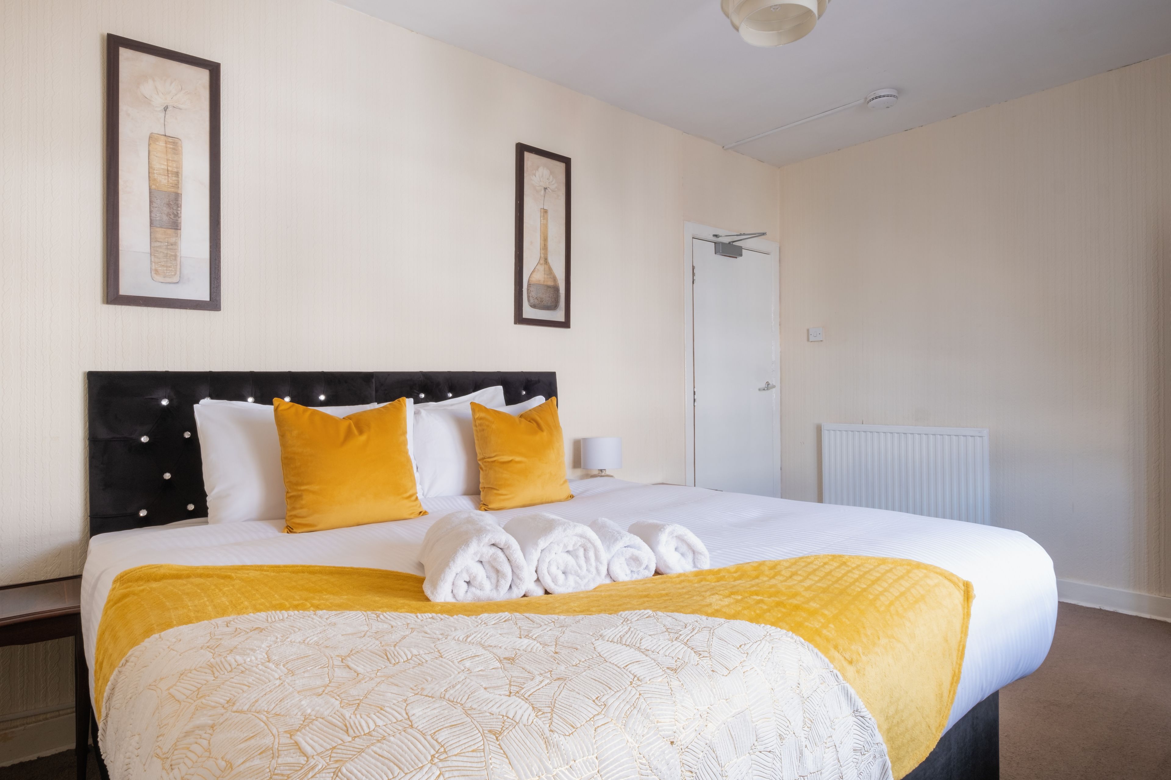 A neatly made bed with a black headboard, white and yellow bedding, and a white wall with two framed pictures.