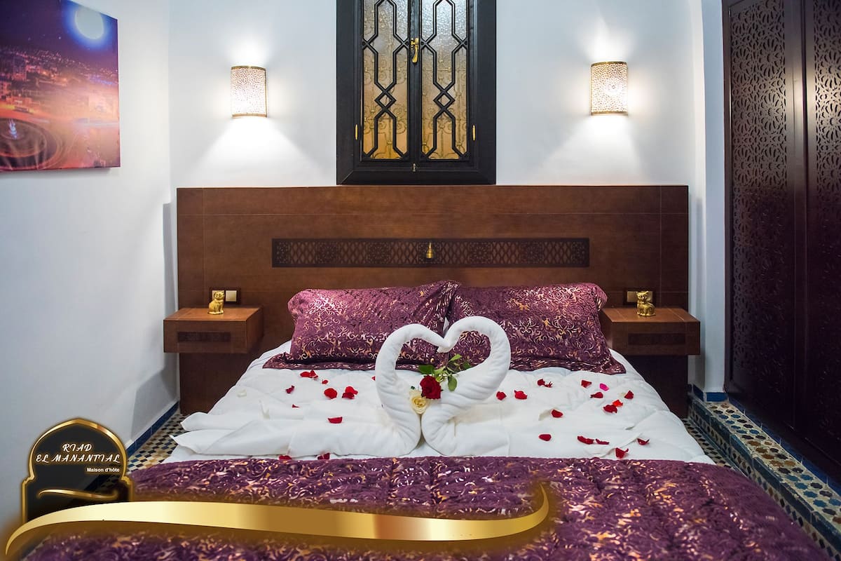 A hotel room with a bed, decorative pillows, a nightstand, a wall-mounted lamp, and a decorative panel.