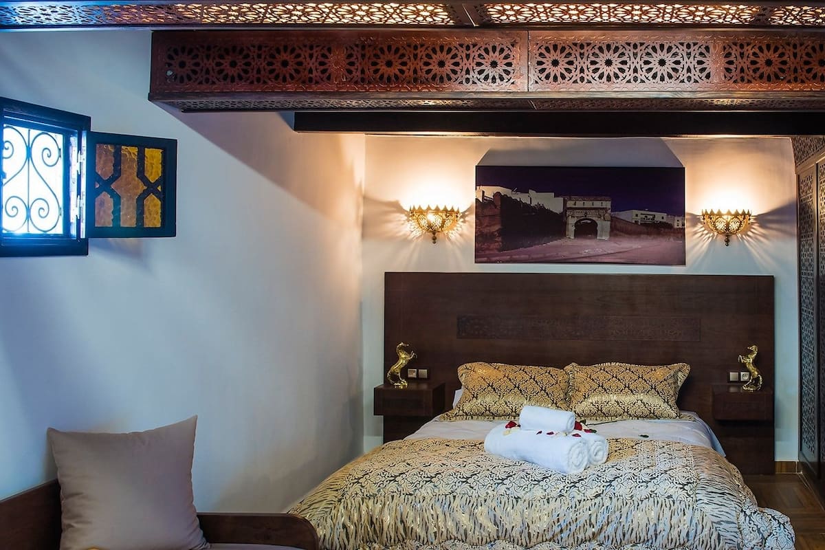 A bedroom with a bed, two nightstands, a headboard, decorative wall panels, and a window with curtains.