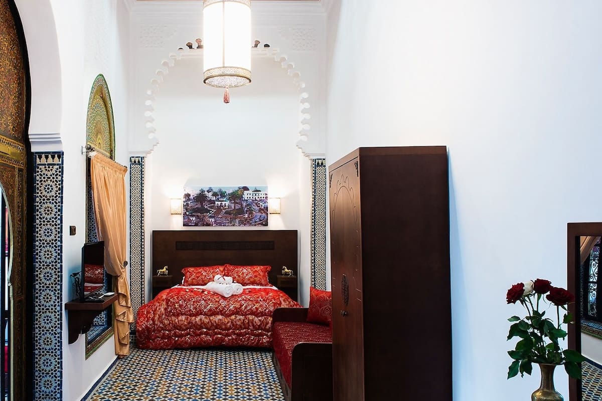 A narrow hallway leading to a bedroom with a bed, a nightstand, a mirror, and a door.