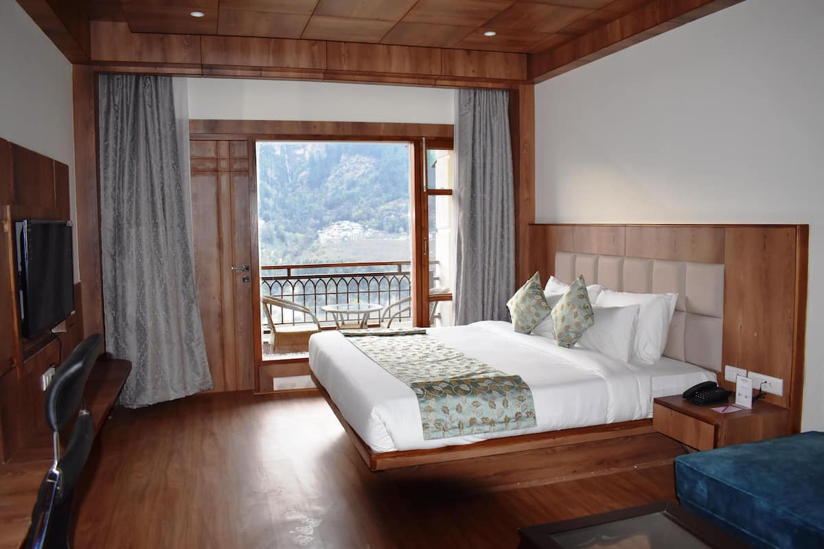 A hotel room with a wooden floor, a large bed with a headboard, a bedside table with a phone, a bench, and a TV.