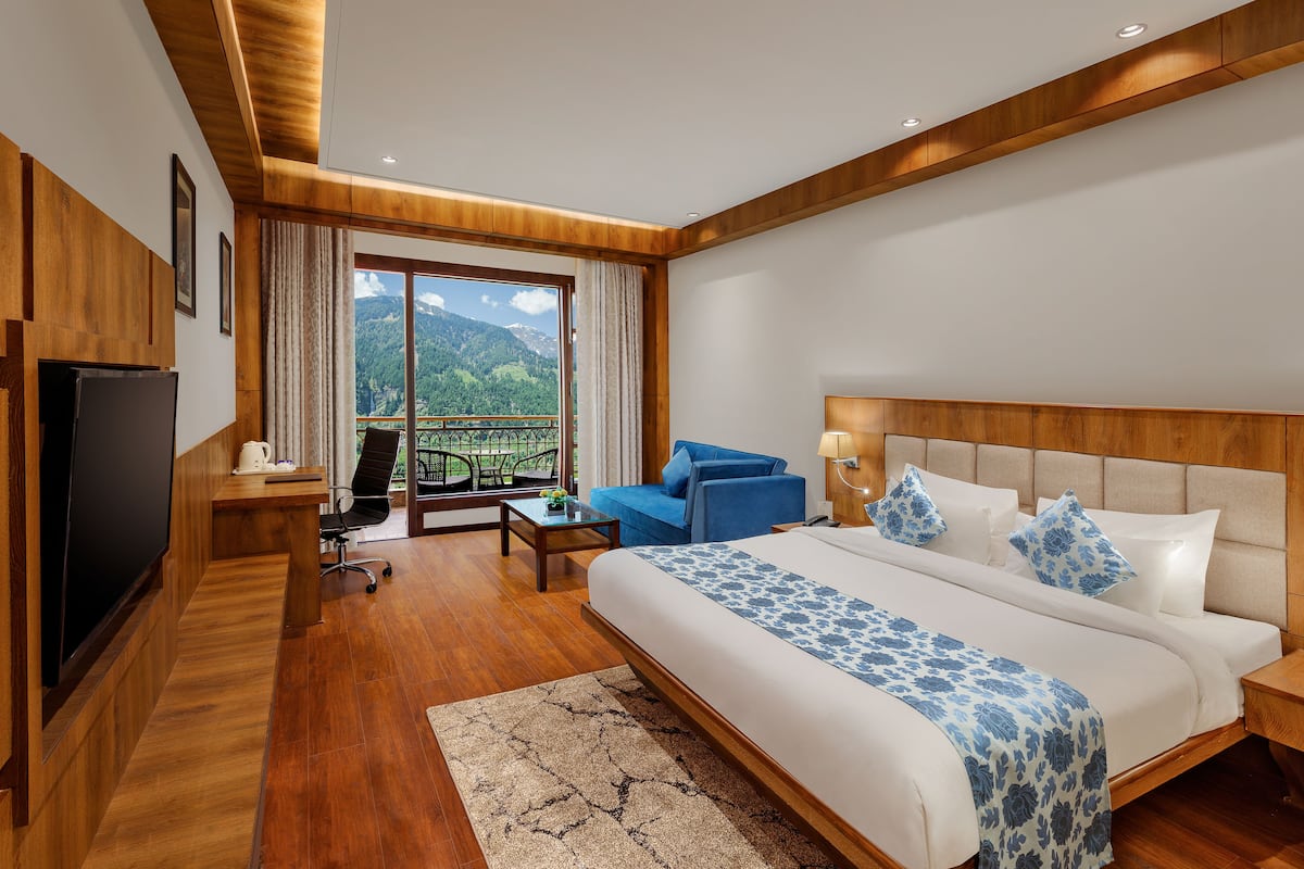 A modern hotel room with a large bed, a TV, a balcony with a mountain view, and wooden flooring.