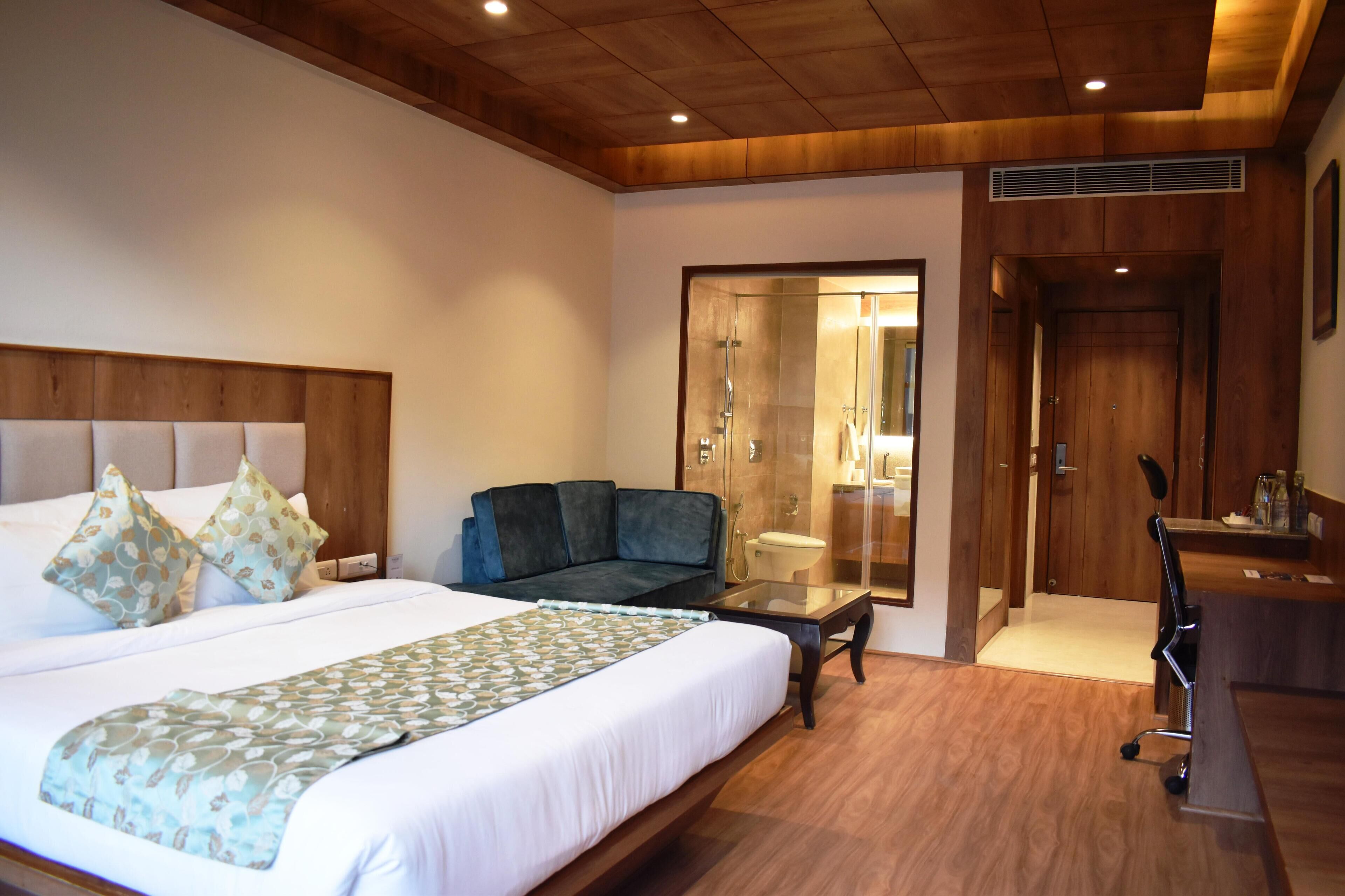 Habitación de hotel con una cama grande, un sofá, un escritorio y un baño visible a través de una puerta de cristal.