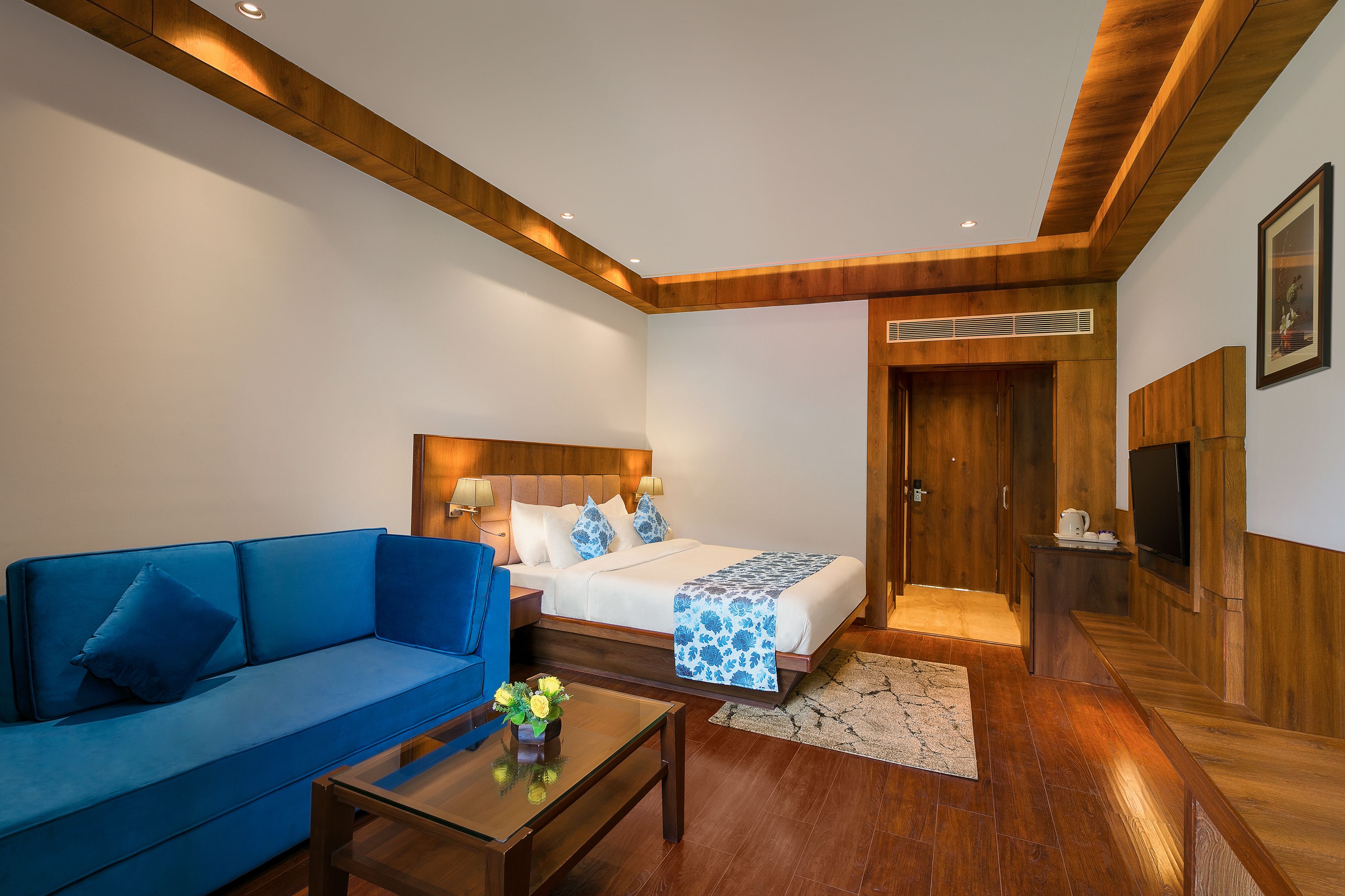 Una habitación de hotel moderna con techo de madera, cama con colcha azul estampada con flores, sofá azul y una mesa de centro con un florero.