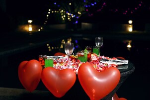 A table set with heart-shaped balloons, glasses, and a drink, with a pool and lights in the background.