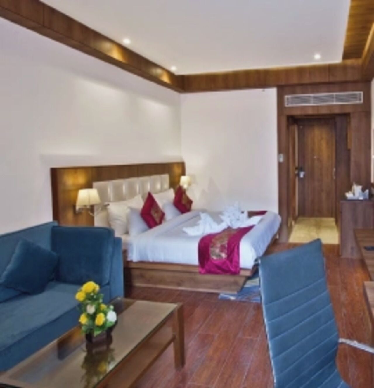 A hotel room with a bed, a blue sofa, a wooden coffee table, and a small table with flowers.