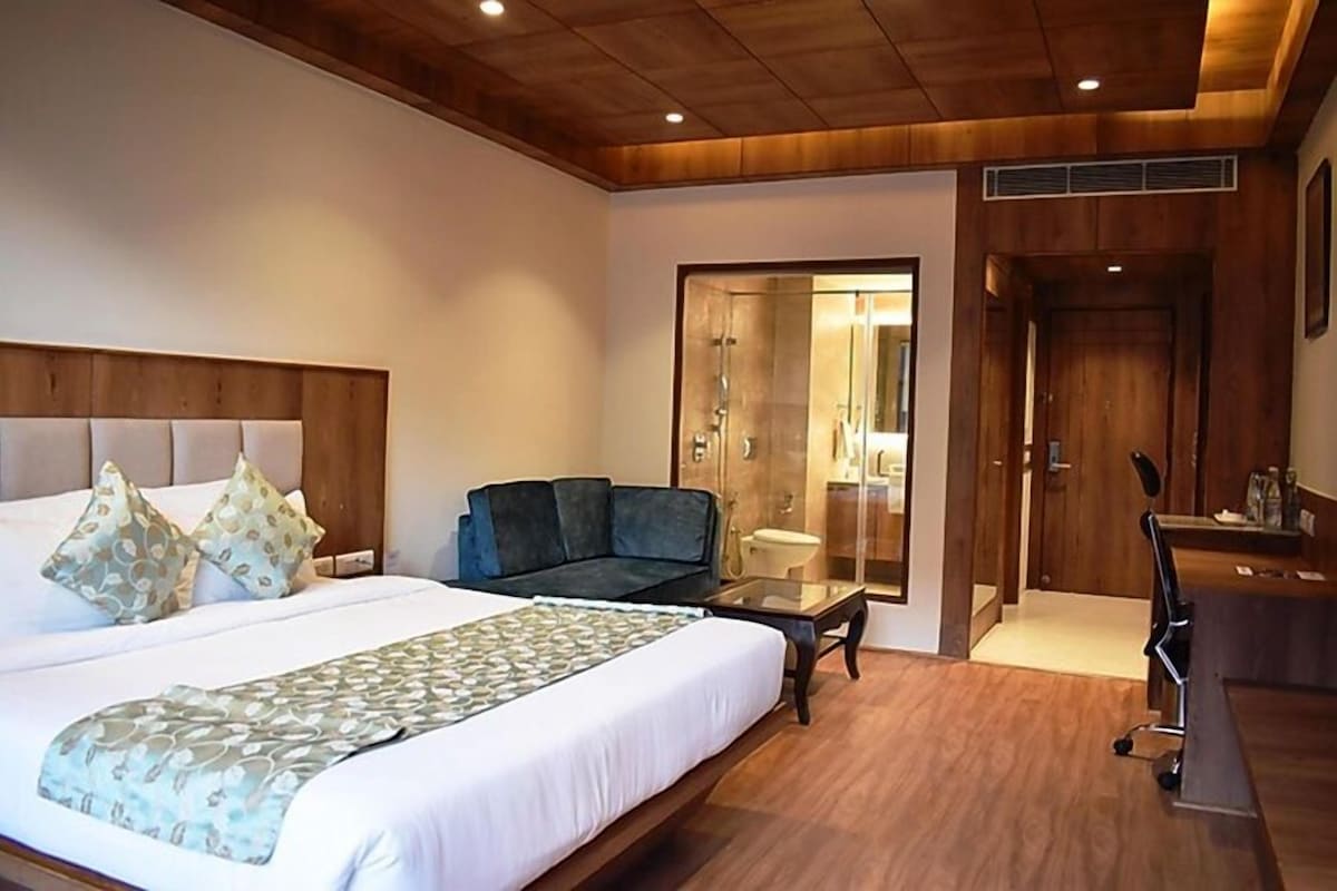 Habitación de hotel con una cama grande, un sofá, un escritorio y un baño visible a través de una puerta de vidrio.