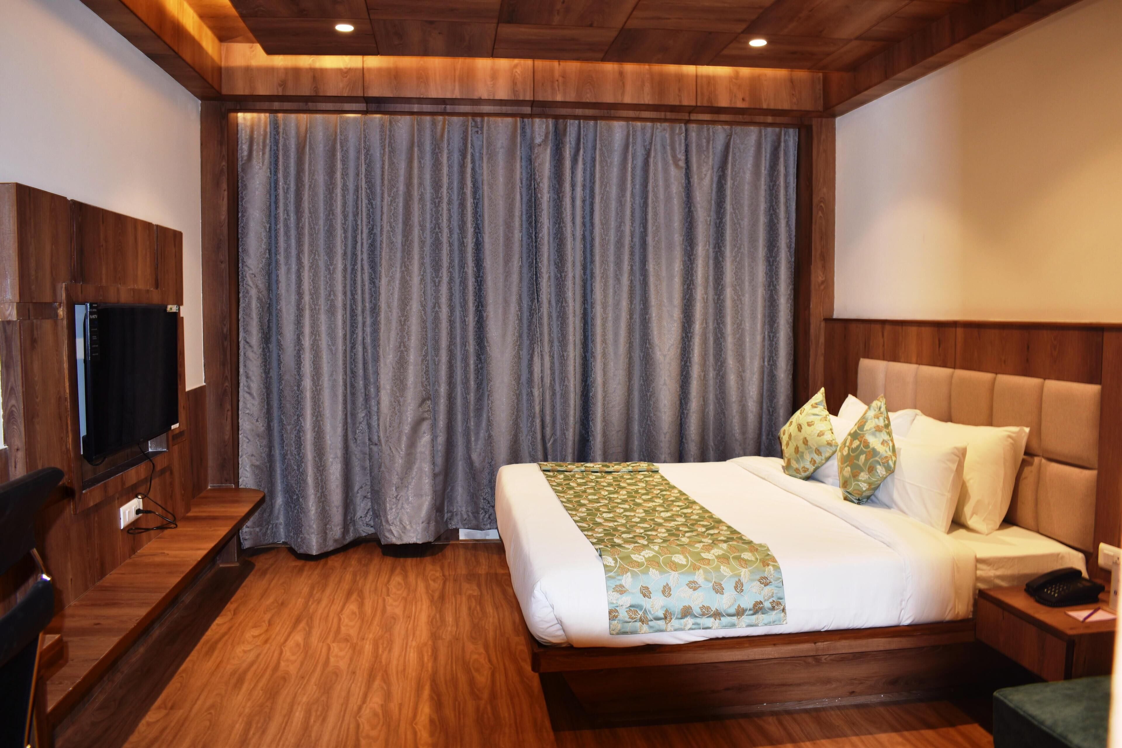 A hotel room with a wooden floor, a large bed with a patterned bedspread, a television on a wooden cabinet, and a large window with curtains.
