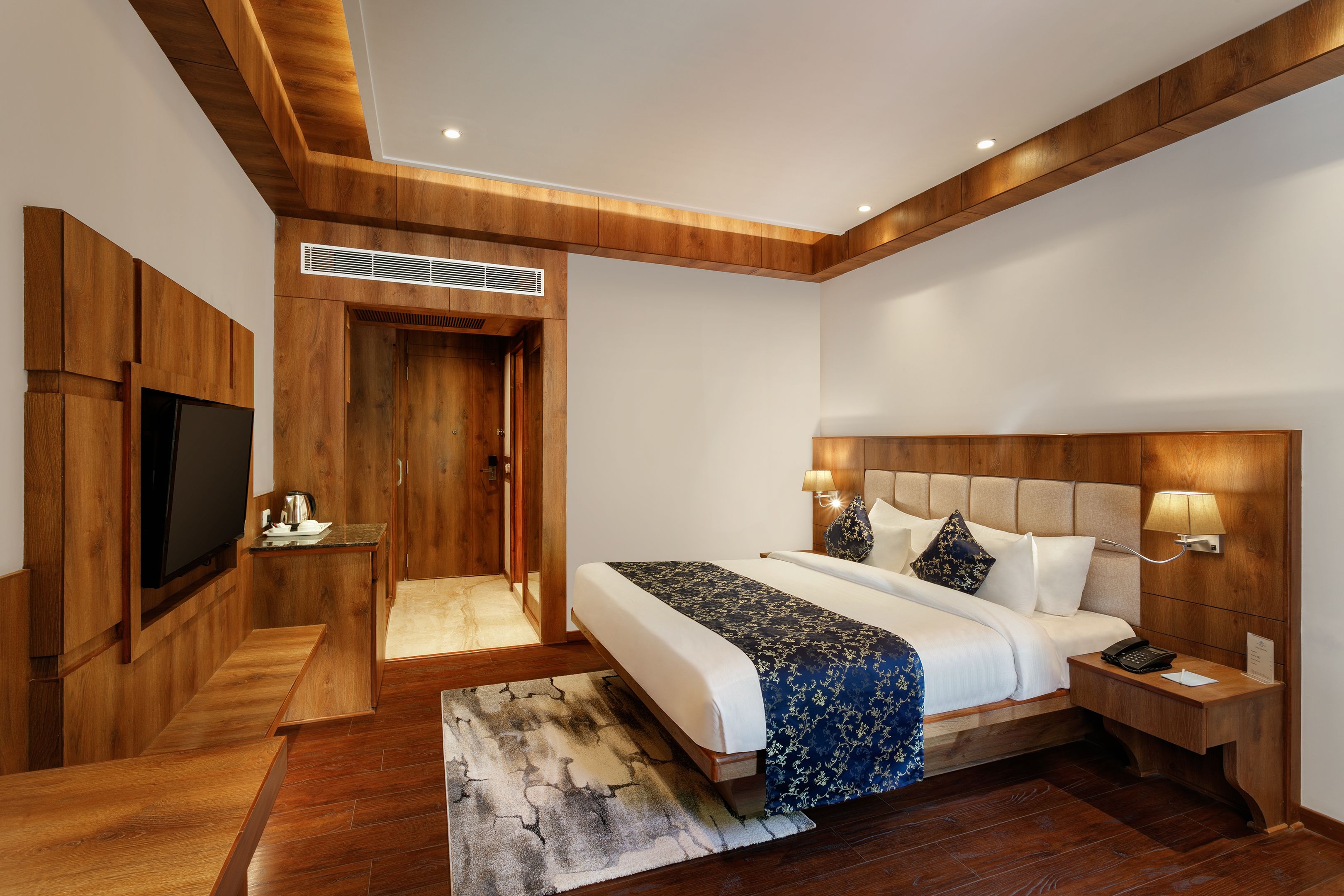 Habitación de hotel moderna con techo de madera, una cama grande con colcha azul y blanca, un televisor de pantalla plana montado en la pared y una mesita de noche con lámpara.