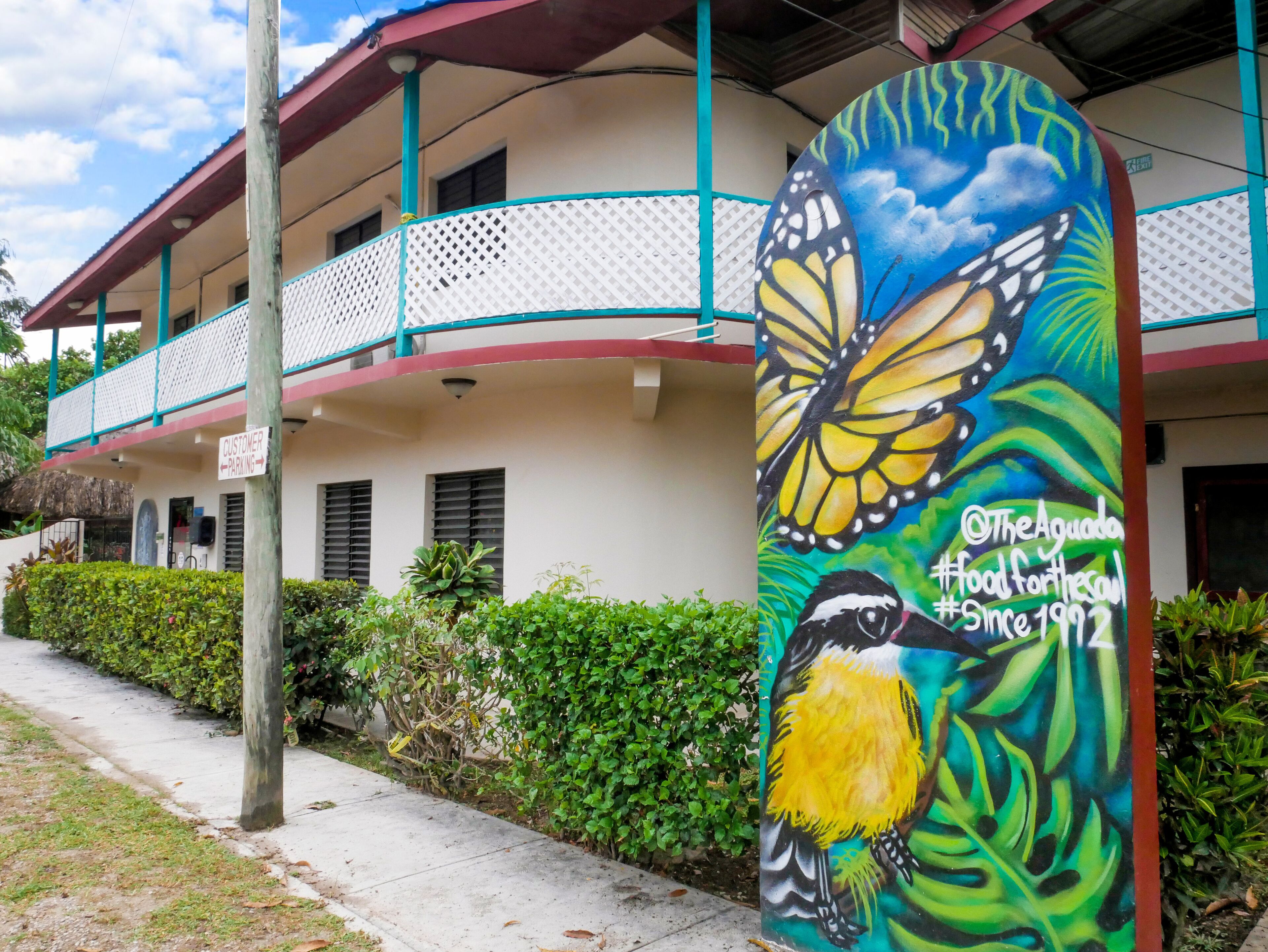 Un edificio con balcón, un cartel que indica estacionamiento para clientes y un colorido mural con una mariposa y un pájaro.