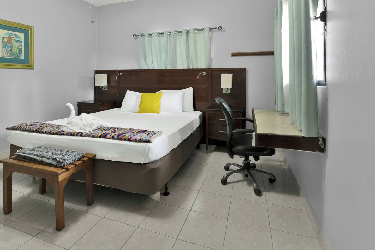 Habitación de hotel con cama, escritorio, silla, una mesita y un cuadro en la pared.