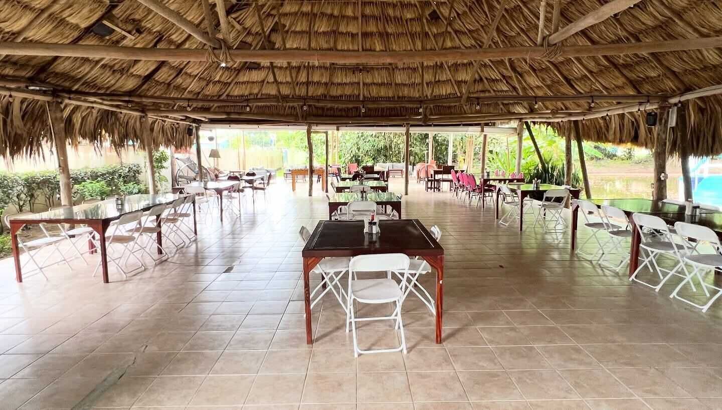 An open-air dining area with thatched roof, tiled flooring, and white chairs. Tables are arranged for dining.