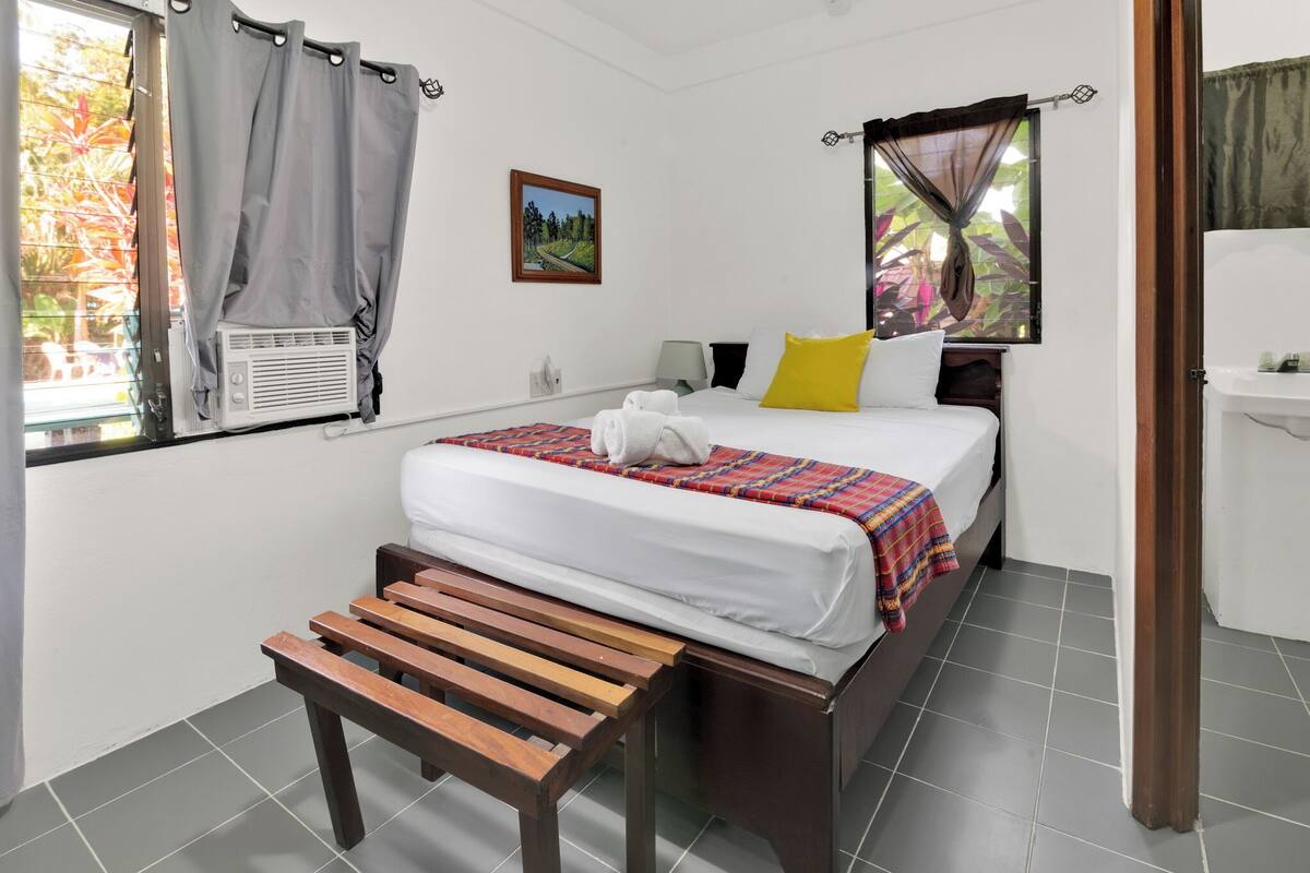 Una habitación de hotel con una cama, un banco, una ventana con cortinas, un cuadro en la pared y un lavamanos.