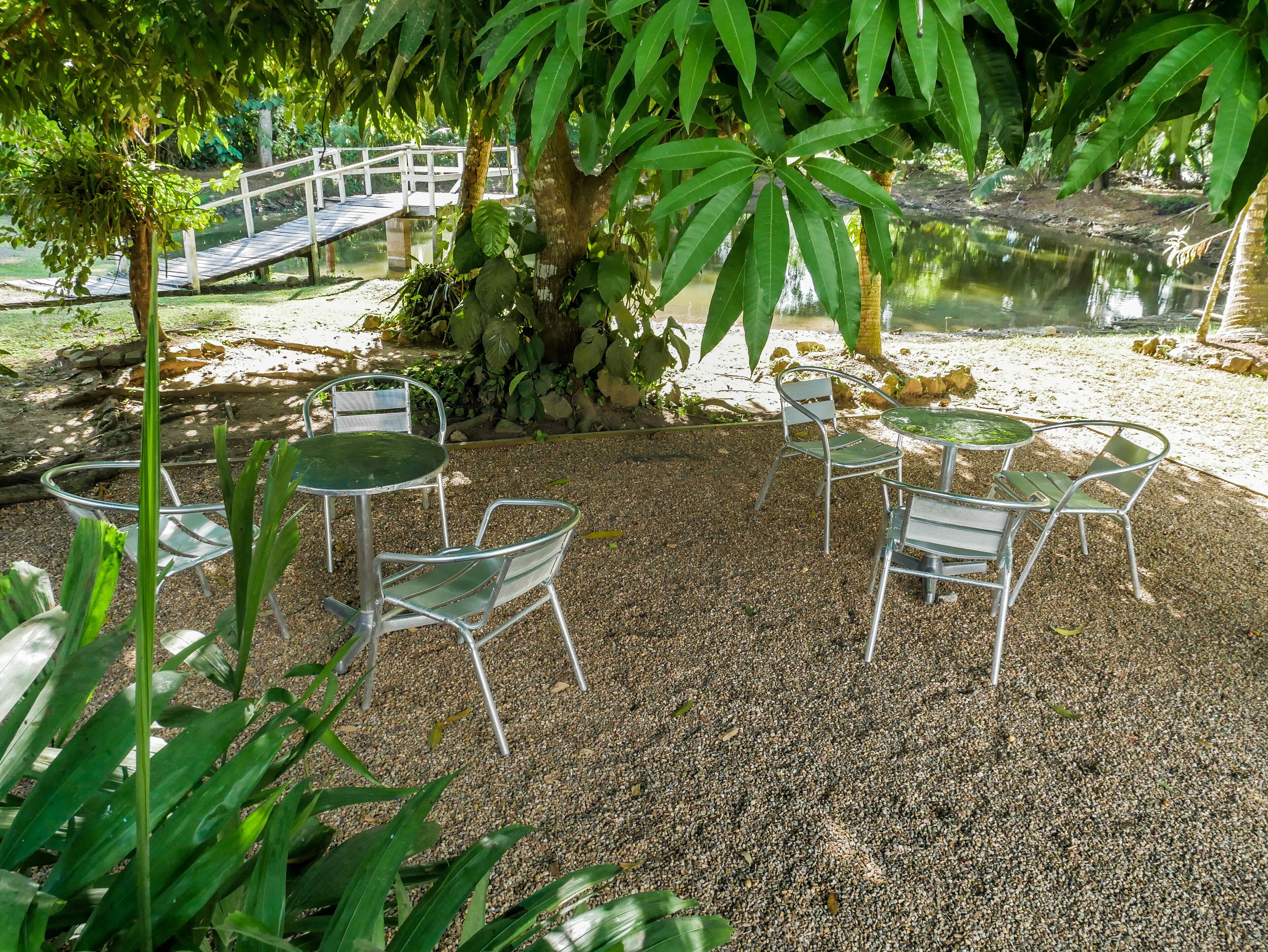 A serene outdoor seating area with metal chairs and tables, surrounded by gravel and lush greenery, with a wooden bridge over a pond in the background.