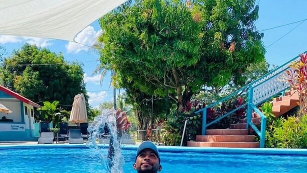 Una piscina con una persona en el agua, una mesa con comida y bebidas, y una escalera que lleva a un edificio.