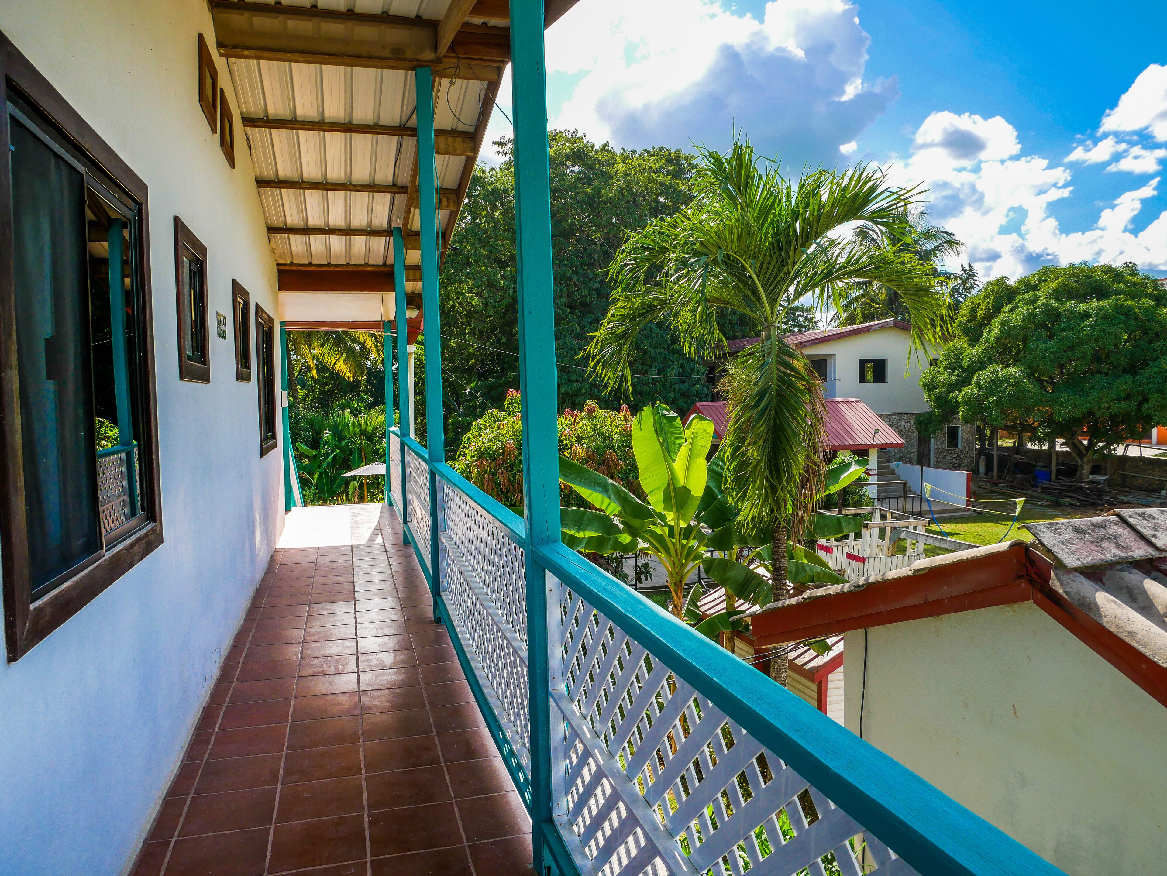 A covered balcony with a view of a garden and a building with a red roof.