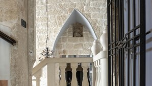Ein Korridor mit Steinwänden und einem spiegelnden Boden, der zu einem Durchgang mit weißem Geländer und schwarzen schmiedeeisernen Toren führt.