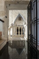 Un pasillo de paredes de piedra con piso reflectante, que conduce a una entrada con puerta de hierro negro y barandilla blanca.