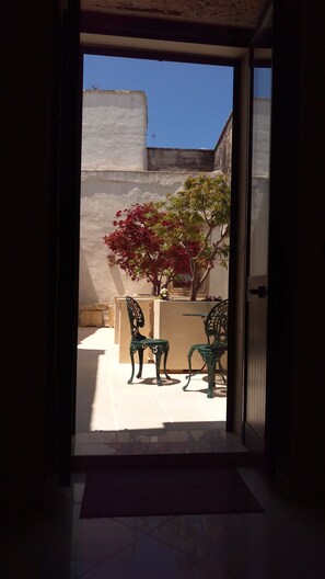 An open doorway leading to a patio with two green metal chairs and a small table, a white wall, and a tree with red leaves. - Palazzo Muro Leccese Relais de Charme (Muro Leccese)