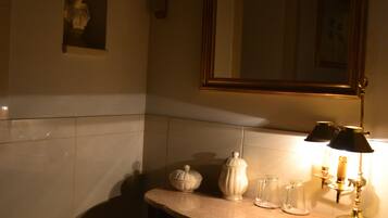Une salle de bain faiblement éclairée, avec un lavabo en marbre, un miroir, un luminaire fixé au mur et une petite statue.