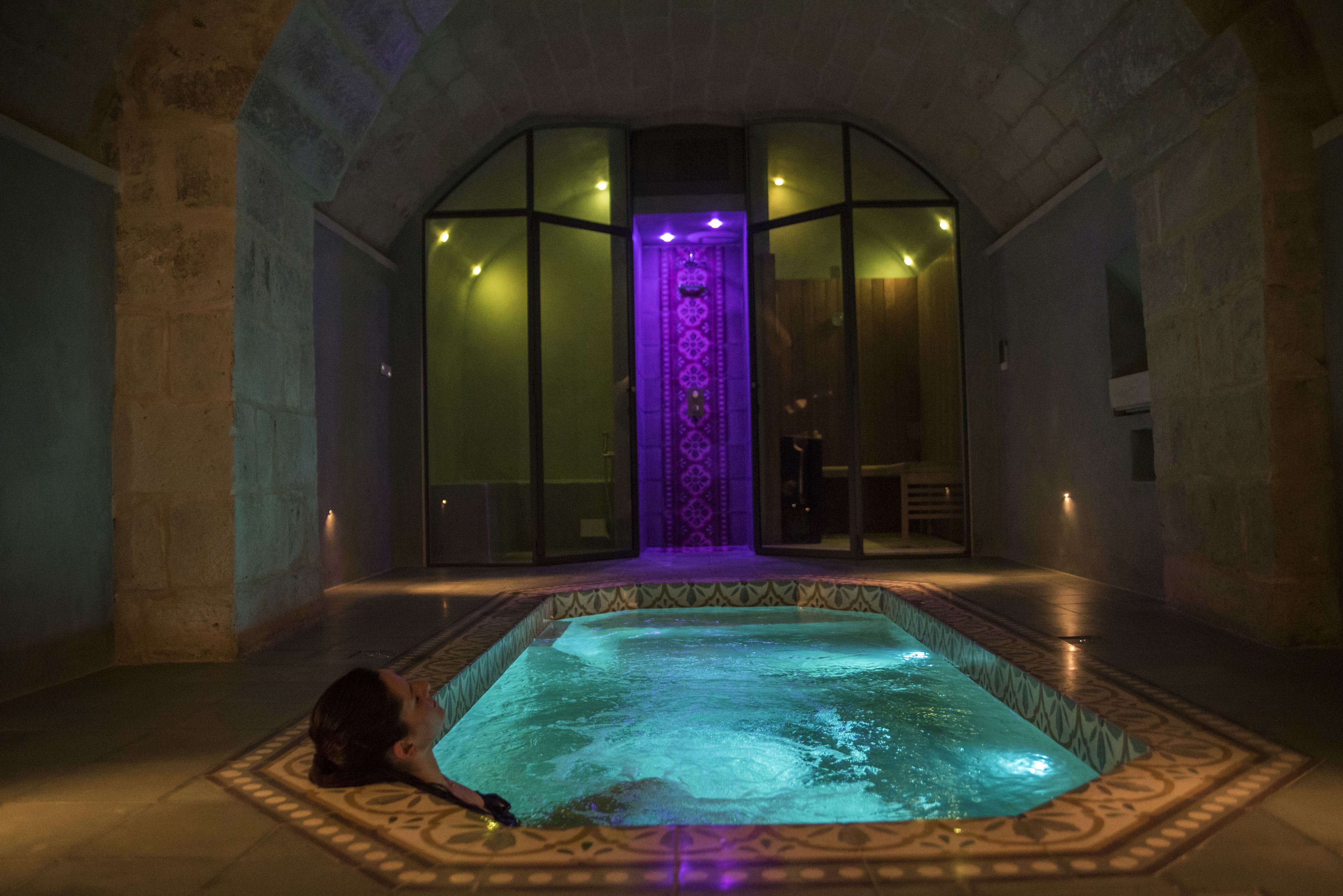 A person relaxing in a hot tub with a patterned floor and a purple-lit wall behind them.