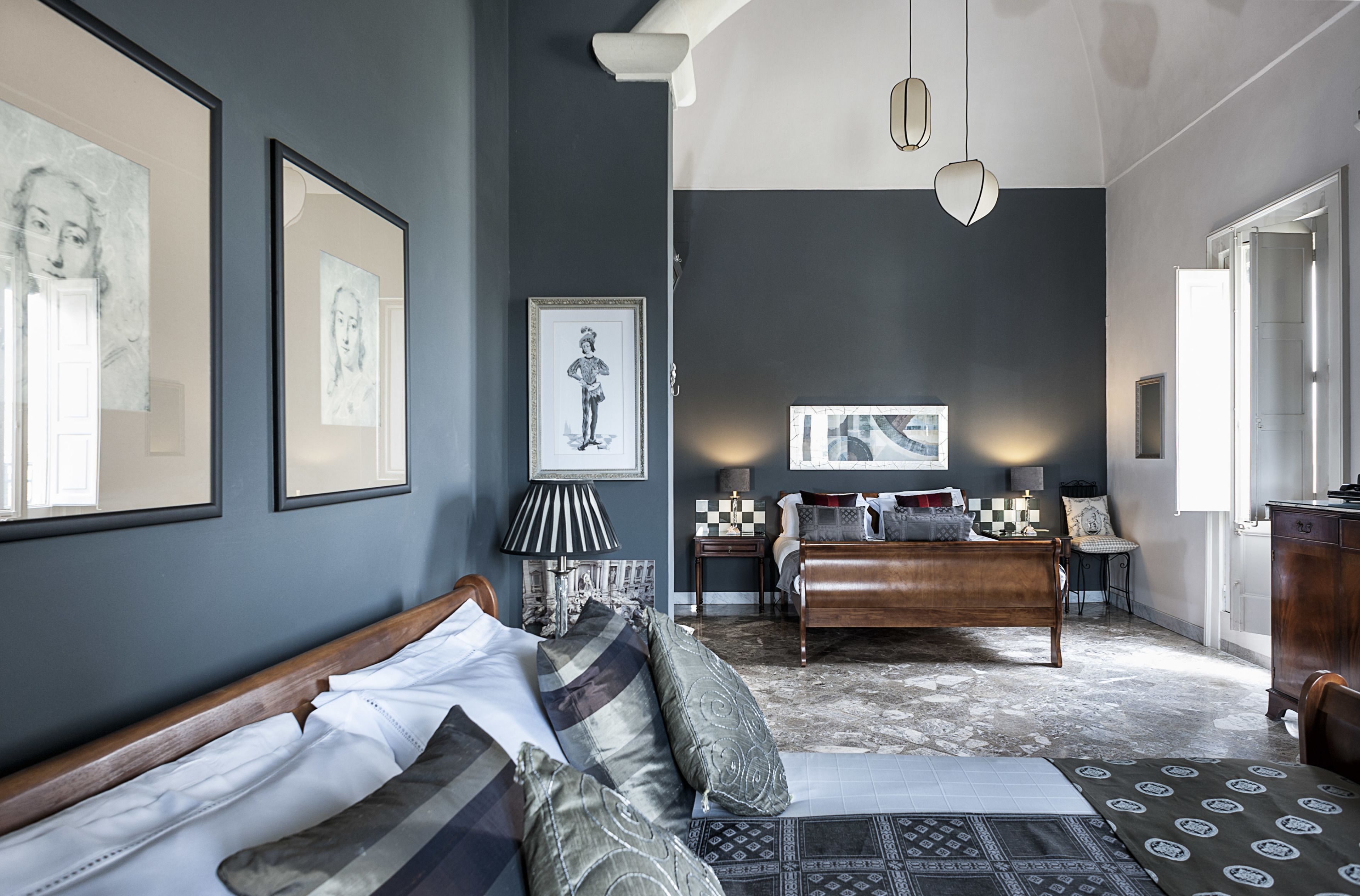 A bedroom with a bed, a sofa, a desk, and a chair. There are framed pictures on the wall and a lamp on the desk.