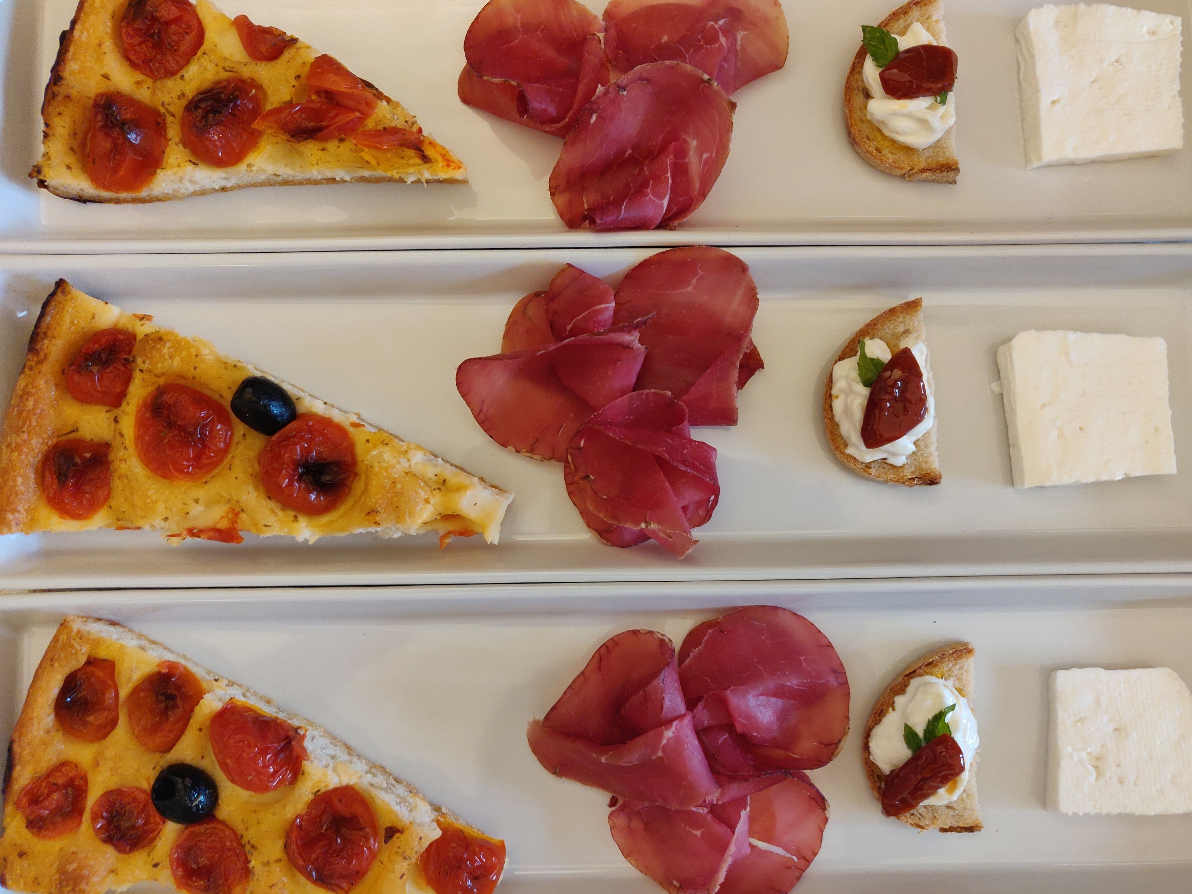 Variedad de alimentos, entre ellos porciones de pizza, prosciutto y queso, sobre un plato blanco.