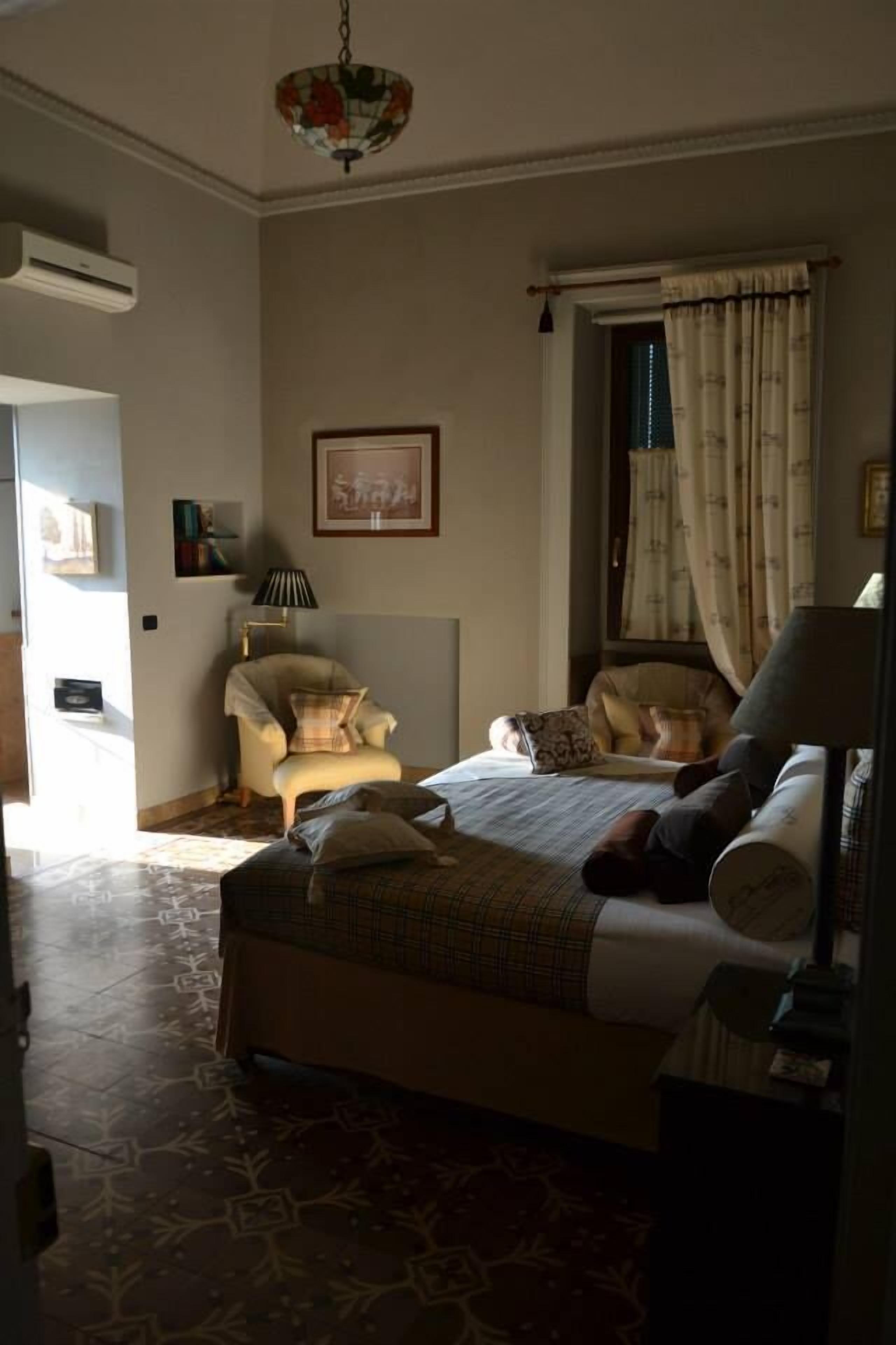 Ein Schlafzimmer mit Bett, Sessel, Lampe und einem Fenster mit Vorhängen.
