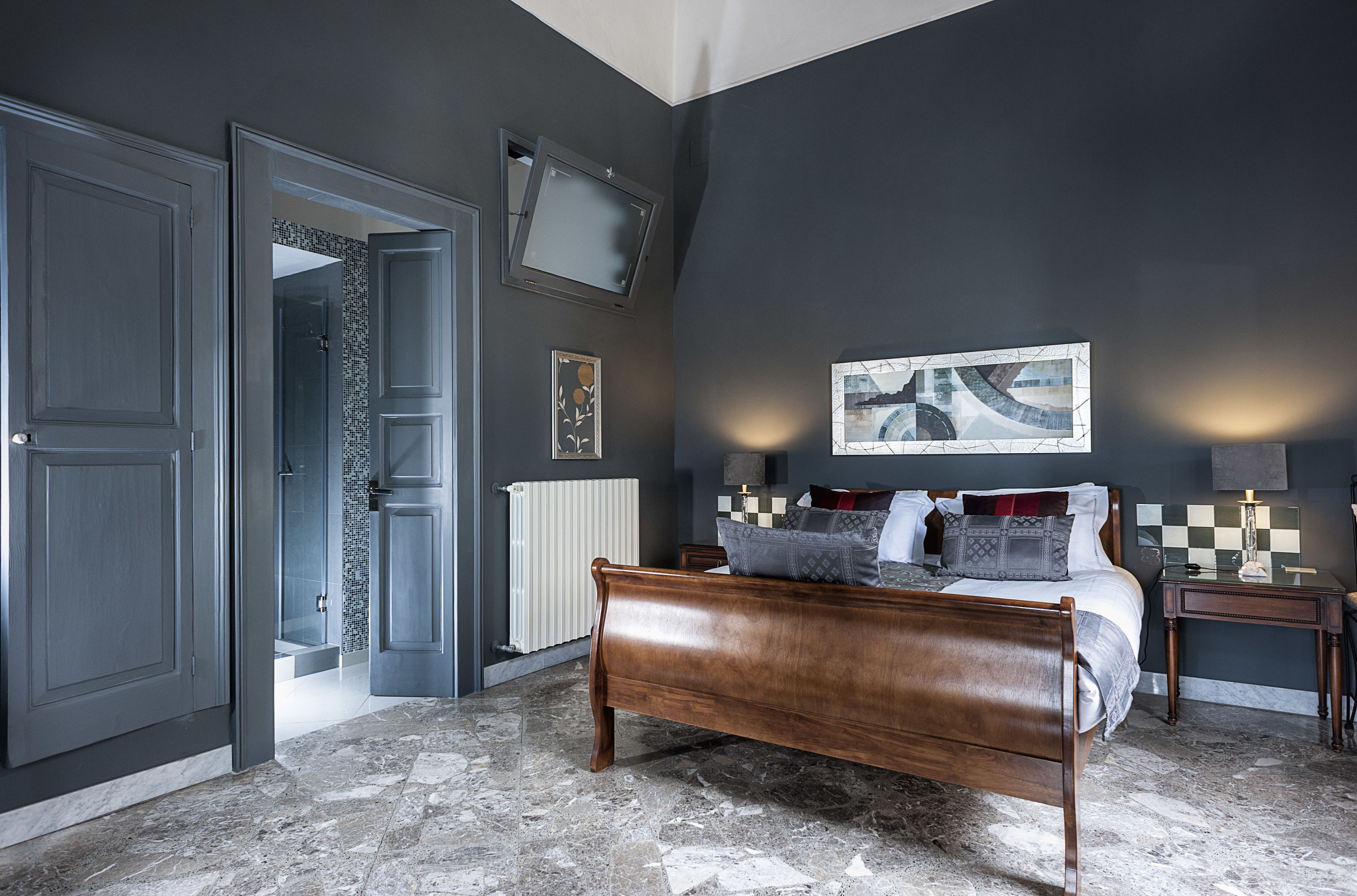 A bedroom with a large bed, a nightstand, a wall-mounted TV, and a door leading to another room.