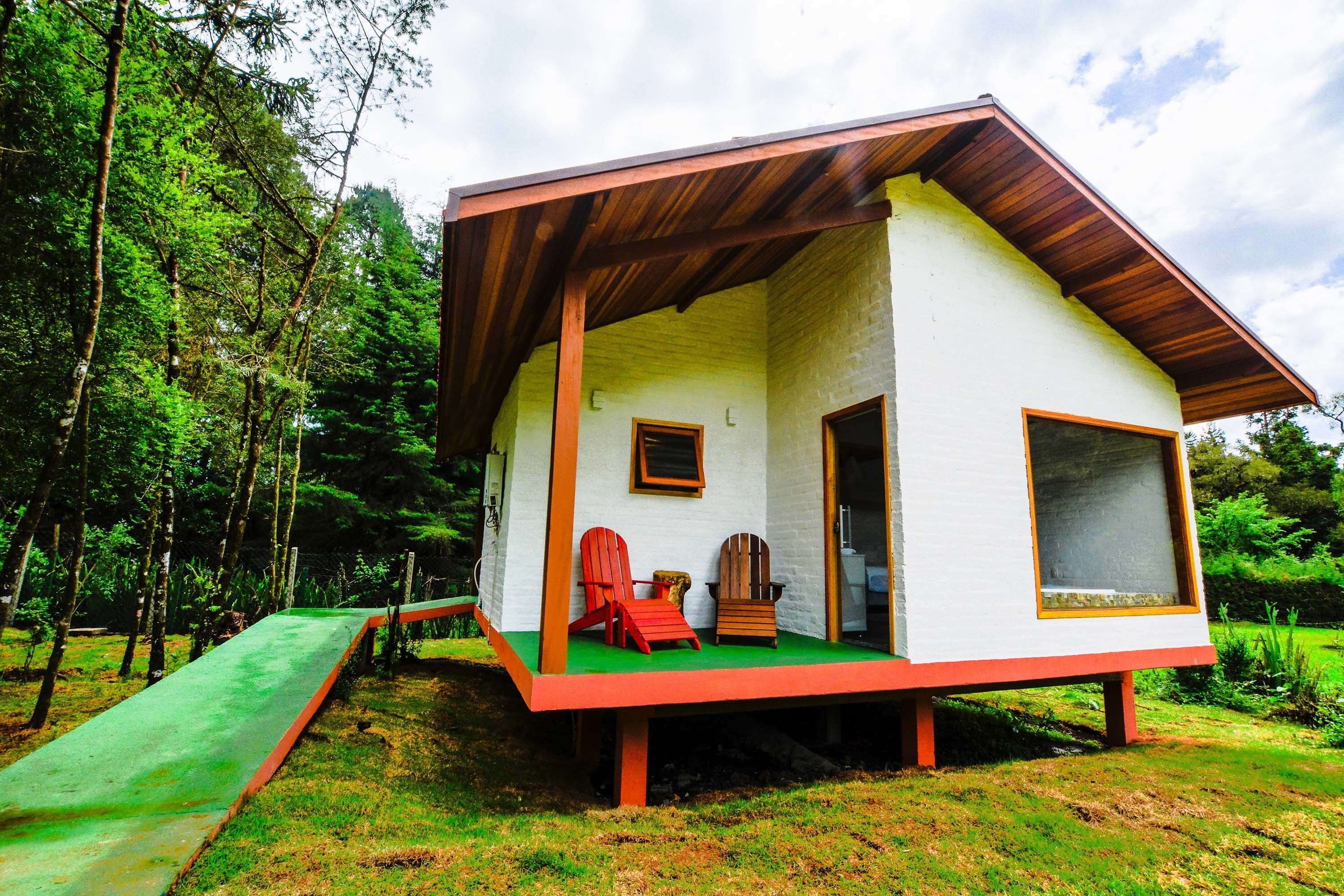 A small cabin with a green ramp leading to a porch, two Adirondack chairs, and a window.
