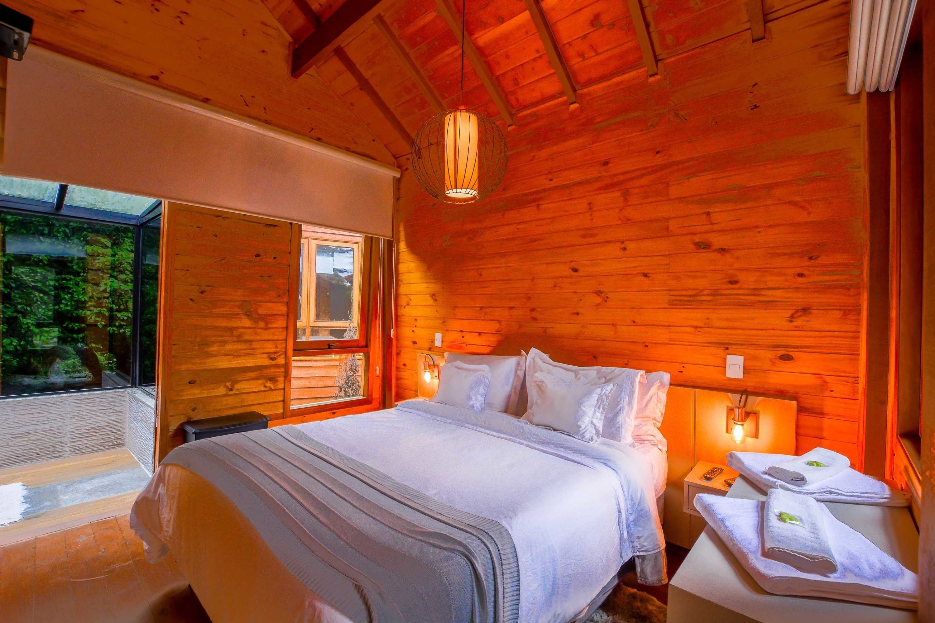 Ein Schlafzimmer einer Holzhütte mit Bett, Nachttischen und einem Fenster mit Blick auf Grünflächen.