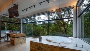 A modern bathroom with a stone sink, a large bathtub, and a view of the forest through large windows. - Resort Magnífico (Camanducaia)