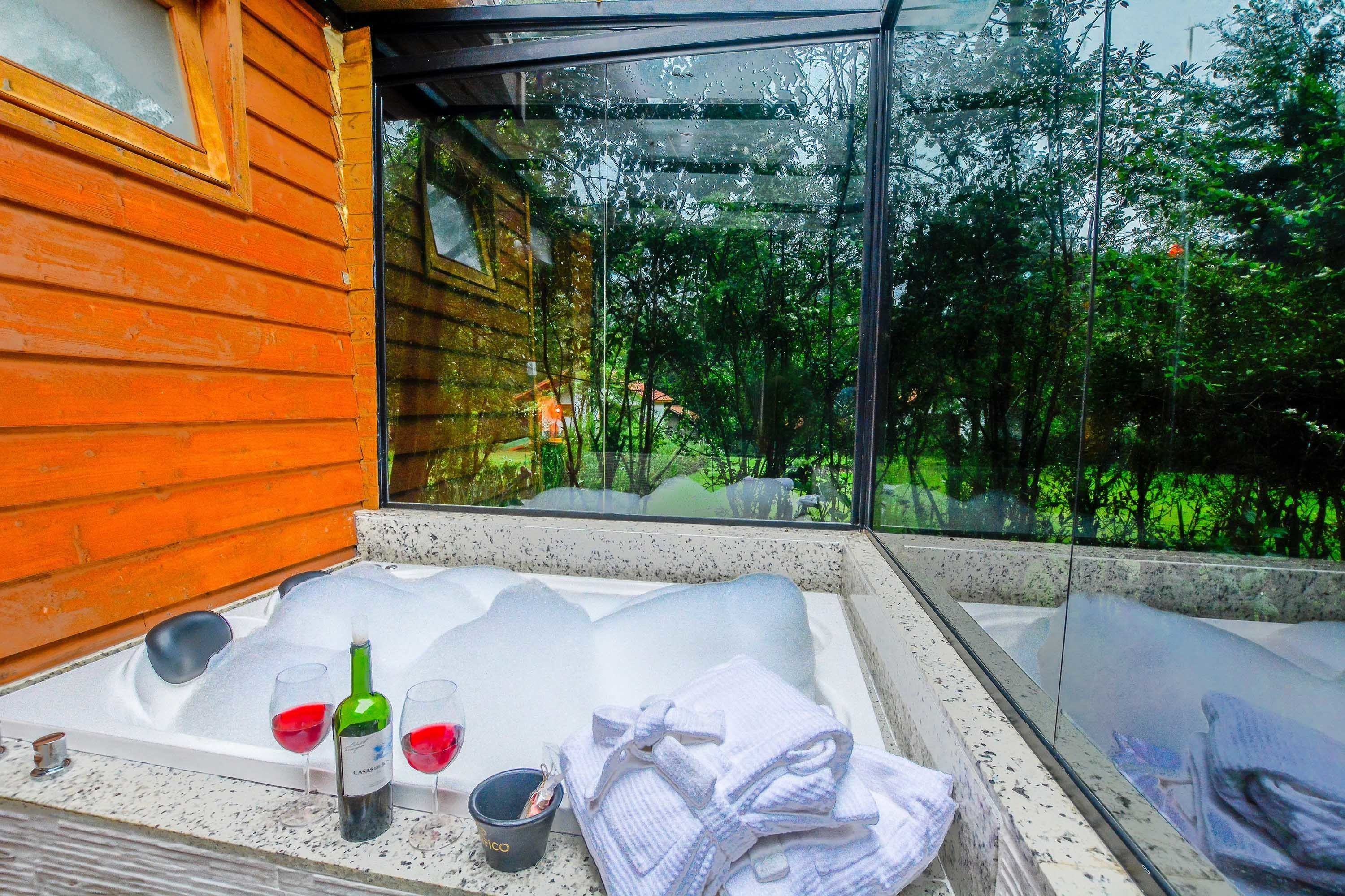 Una habitación con paredes de vidrio y fachada de madera, incluye una bañera y una mesa con copas de vino y una botella.