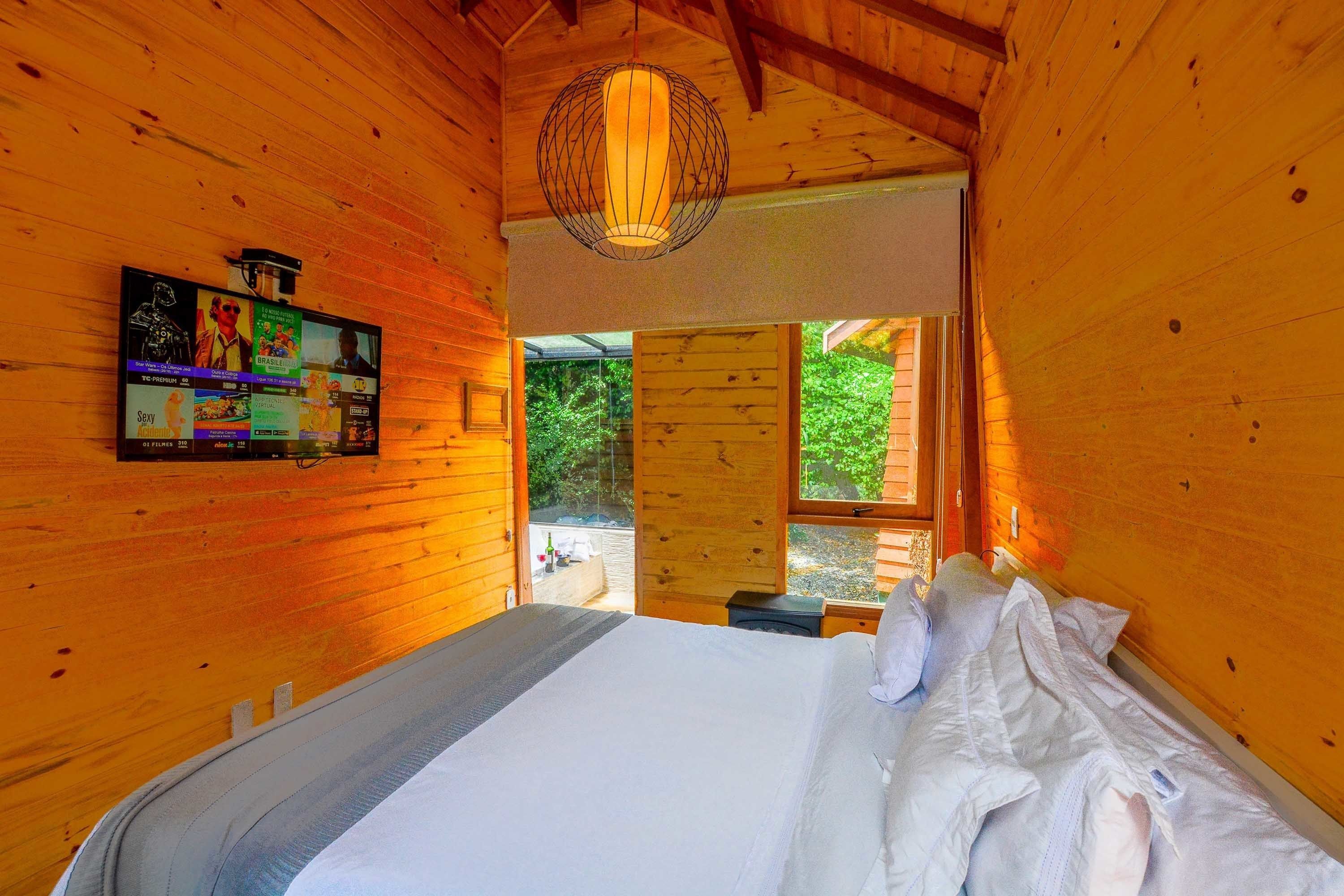 A wooden cabin bedroom with a bed, a wall-mounted TV, and a large window.