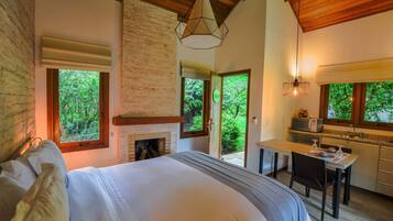 A bedroom with a large bed, a fireplace, a dining area, and a kitchen with a view of the forest.