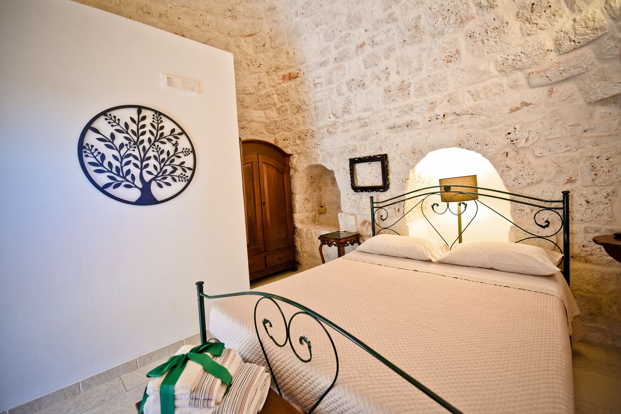 Un dormitorio con pared de piedra, una cama con estructura metálica, un armario de madera y un cuadro colgado en la pared con un árbol.